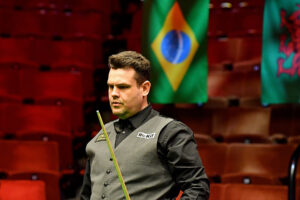 Igor Figueiredo in the Crucible Theatre arena at the 2021 World Seniors Snooker Championship. Behind him is a Brazilian flag hanging above him and empty seats as the event was played behind closed doors due to Covid.