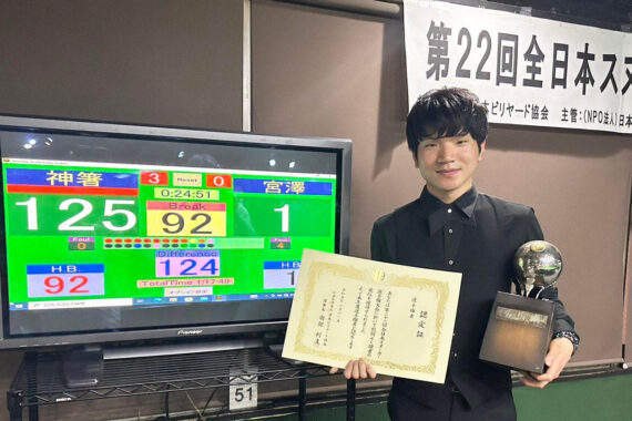 Keishin Kamahashi poses with the trophy in front of the television scoreboard. He also holds a certificate.