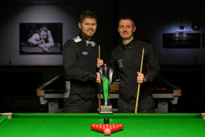Daniel Womersley and Michael shake hands with the Q Tour trophy on the table just in front of them. Both players are in waistcoats, bow-ties, shirts and trousers.