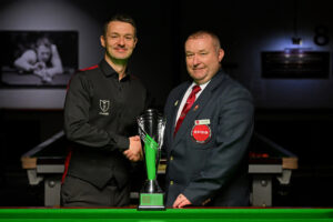 Michael Holt and tournament director Stuart Barker shake hands with the Q Tour on the table just in front of them.
