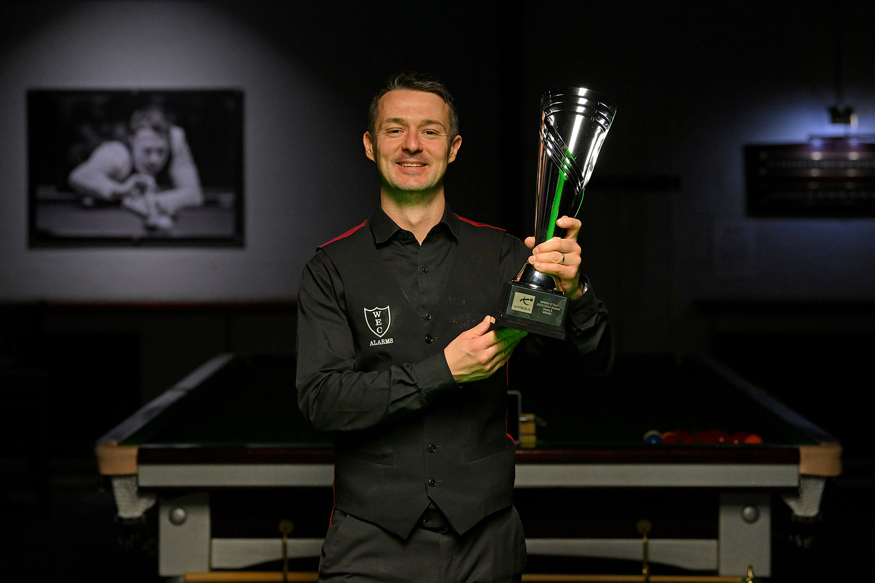 Michael Holt in his waistcoat, shirt and trousers lifts the Q Tour trophy in front of a snooker table.