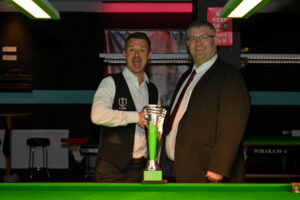 A very happy Michael Holt shakes hands with tournament director James Chambers at the table with his trophy.