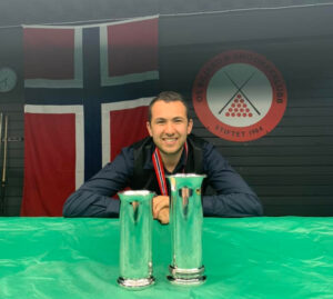 Nassim Sekat poses by crouching down with forearms on the covered up table and trophies in front of him and with medal on.