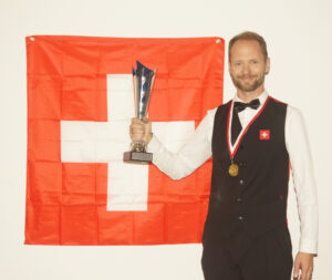 A smiling Risto Värynen wears his medal and holds his trophy aloft in front of the Swiss flag.
