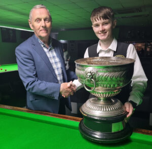 Robbie McGuigan shakes hand with the tournament sponsor and in front of the Northern Ireland Championship trophy on the table.