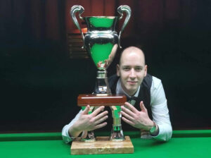 Zsolt Fenyvesi leans on a snooker table with his trophy shows five fingers on both hands to signal his 10 wins in the event.